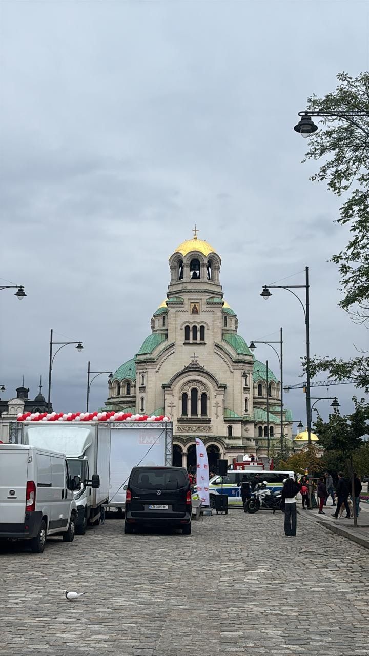 A cathedral with a gold central dome and green domes at the end of a cobbled street, with parked vans and lamp posts under an overcast sky