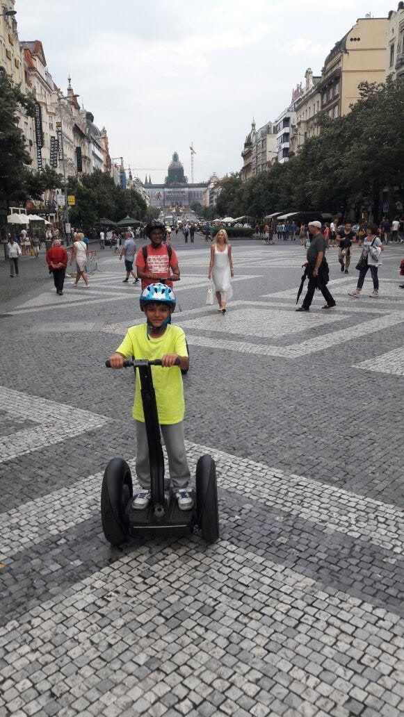 A child on a two-wheeled personal transporter in a wide pedestrian square, with a domed building at the far end